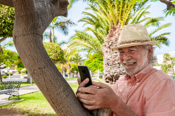 istock Smiling attractive retired man with beard and hat using smart phone standing in a public park between meadow and plants - active lifestyle for a pensioner people 1255968008