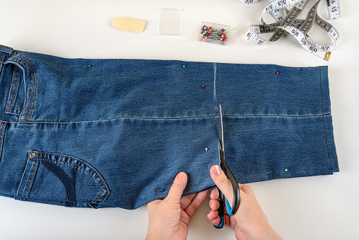 Caucasian woman hands cutting out a folded in half blue denim capris with scissors on a white table. Shorten the jeans with scissors and sewing pin. DIY summer clothes. Top view.