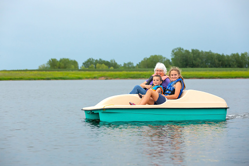Close-up of a young boy and girl in the paddleboat with Grandma. They are floating on a small lake on an overcast summer day. All three are smiling at the camera.