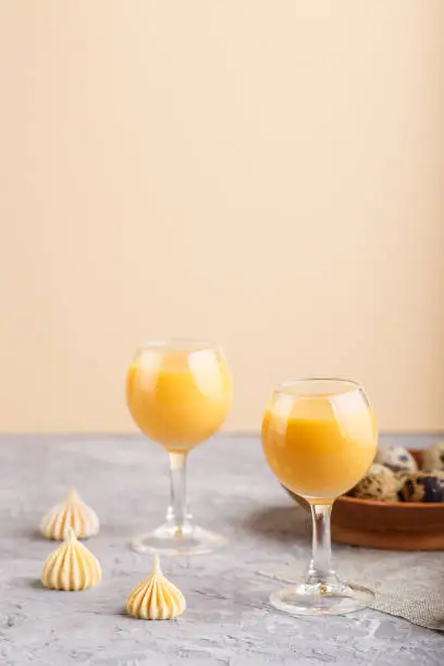 Sweet egg liqueur in glass with quail eggs and meringues on a gray and orange background. Side view, copy space.