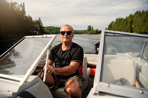 50 + man driving small boat on a lake. He is wearing hat and  black t-shirt over swimwear, and is looking at the camera with a smile. Horizontal waist up outdoors shot with copy space.