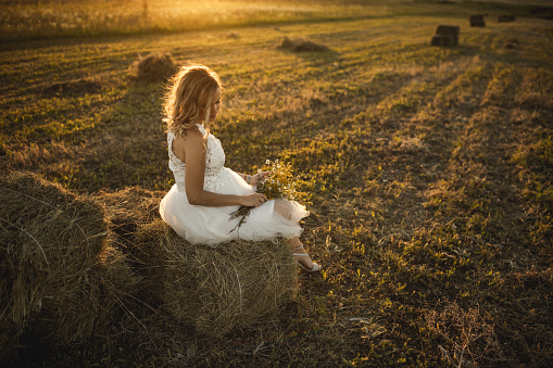 Young blonde bride in wedding dress sitting on hay bale in stubble field and holding wildflower bouquet