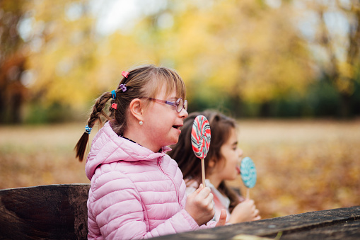 Little girl with Down syndrome and her sister eating lollipops in the park.