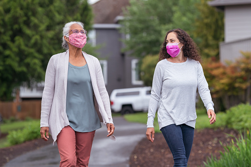 A mixed race senior adult woman is out for a walk with her adult daughter. The two women are wearing protective face masks. They are enjoying the outdoors while taking proper social distancing measures to avoid viral infection of COVID-19.