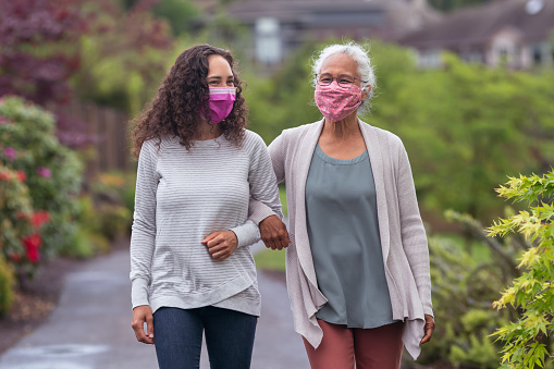 A mixed race senior adult woman is out for a walk with her adult daughter. The two women are wearing protective face masks and are walking with their arms linked. They are enjoying the outdoors and getting some exercise during COVID-19.