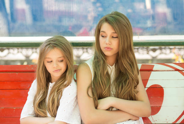 Small sisters offended and upset after quarrel, portrait closeup two siblings back to back looking at each other skeptical outside isitting on a bench a city bus station