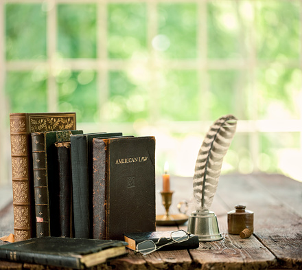 Vintage study area with books, quill pen and candle on an old wood desk in front of a window