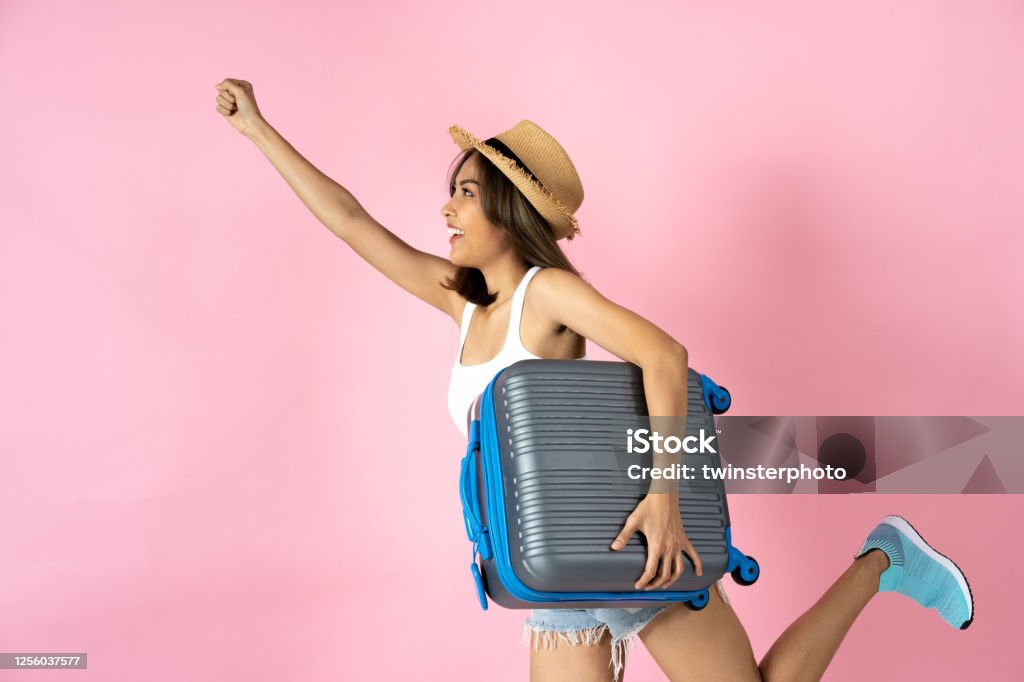 Young woman carrying suitcase with arm raised against pink background Adventurous young woman holding luggage with arm raised against pink background Journey Stock Photo