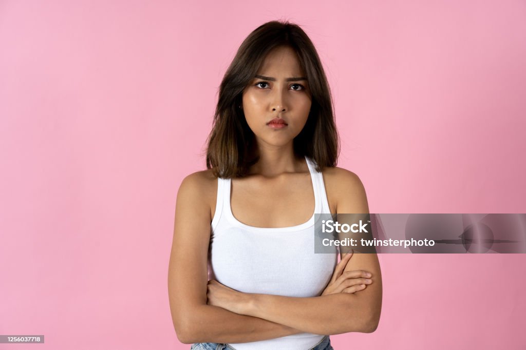 Portrait of young woman with arms folded against pink background Serious young woman crossing arms and looking at camera against a plain pink background Teenager Stock Photo