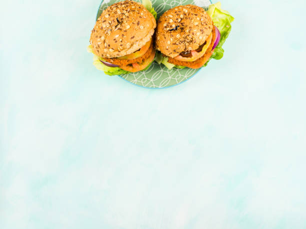 Vegan burgers with soy and vegetable patties, onion, tomatoes and lettuce with spelt and seeds buns on green dish over turquoise background. Copy space, flat lay