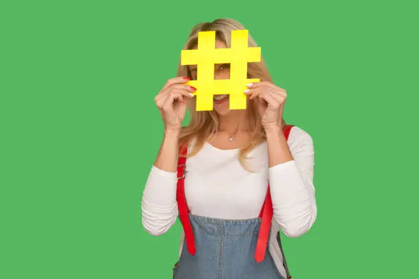 Photo of Blog comments, internet trends. Portrait of positive woman in denim overalls looking through hashtag symbol