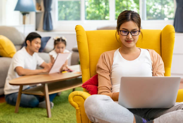 Working mom work from home with dad and daughter in background Asian working mom work from home in living room with background of father and daughter reading tale story book while city lockdown. Diversity concept of working mom and family dad and girl. working at home with children stock pictures, royalty-free photos & images