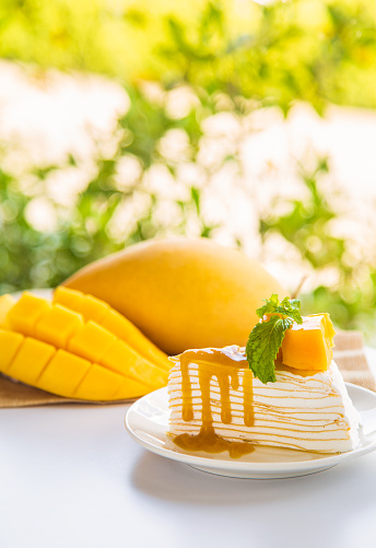 Close up mango crepe cake sliced on a white plate decorated with a piece of mango fruits and peppermint on a wite table with mango fruit and green nature background.