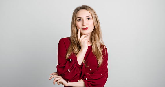 Photo of charismatic Pensive European young woman with long blonde hair keeps hands together near chin, has cute expression, wears red blouse. Copy space for your text