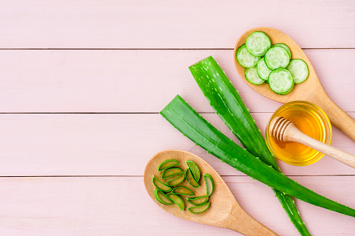 Green fresh aloe vera leaf with cucumber slice in wooden spoon and honey isolated on pink wood table background. Natural herbal medical plant, skincare, health and beauty spa concept. Flat lay.