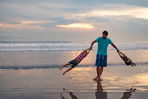 Rear view of father and children, a boy and a girl,  having fun while spinning around on the beach at dusk. Copy space.
