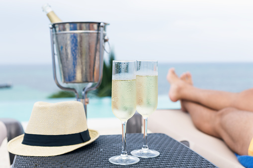 Cold champagne bottle in ice bucket and two glasses of sparkling wine on rattan table near infinity swimming pool