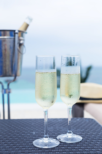 Cold champagne bottle in ice bucket and two glasses of sparkling wine on rattan table near infinity swimming pool