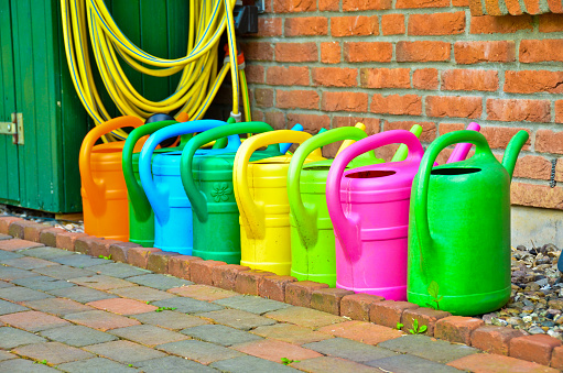Many colourful watering cans standing in a line in front of a wall of red bricks