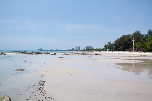Panoramic view along beach of Hua Hin. At beach are some rocks. A few people are on beach. In background are hotels