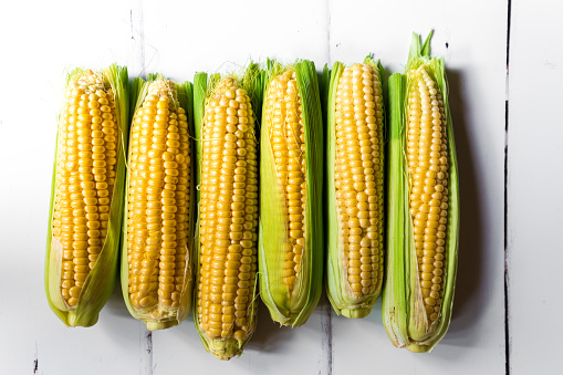 Macro color image depicting fresh corn cobs in a row on a white wooden surface.