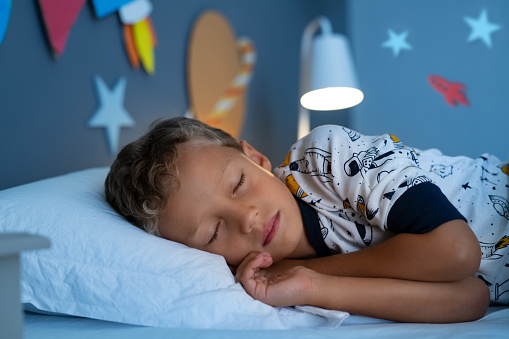 Cute little boy lying on side on bed and sleeping in bedroom decorated with planets and stars. Child sleeping in decorated room and dreams of going into space. Kid sleeps deeply with closed eyes in his space themed room and dreaming of being an astronaut.