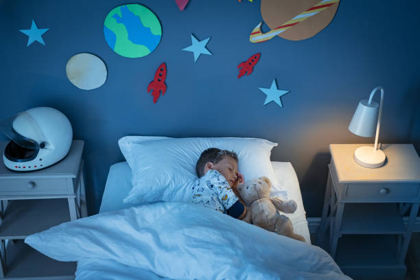 Boy sleeping and dreaming a future in the space High angle view of little boy dreaming of becoming an astronaut while sleeping with teddy bear in space decorated room. Top view of dreamer child sleeping on bed with solar system and planet decoration during the night. Cute kid with astronaut helmet on side table and the light on. science and technology kids stock pictures, royalty-free photos & images