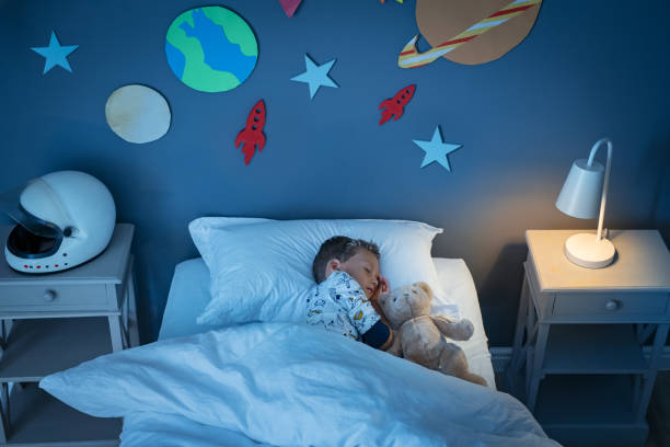 Boy sleeping and dreaming a future in the space High angle view of little boy dreaming of becoming an astronaut while sleeping with teddy bear in space decorated room. Top view of dreamer child sleeping on bed with solar system and planet decoration during the night. Cute kid with astronaut helmet on side table and the light on. one little boy stock pictures, royalty-free photos & images