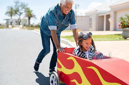 Senior grandfather helping grandson ride on kids toy car. Happy old man pushing car while boy driving on street. Grandparent playing with little boy on gokart.