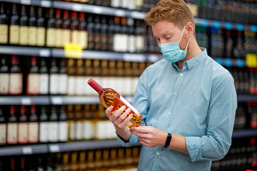 Young man wearing protective face mask in supermarket, holding a bottle of rose wine and looking at label, COVID-19 pandemic era