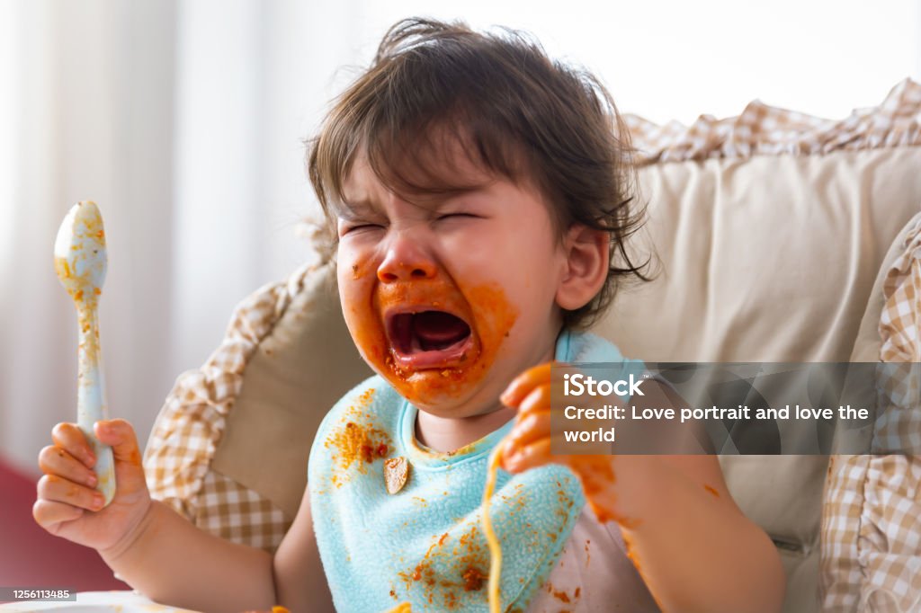 Adorable little toddler girl or infant baby crying when unsatisfied when finished eating food on baby chair. Cute infant girl get hungry and want more food. Mix race daughter get dirty Kid get tantrum Child Stock Photo