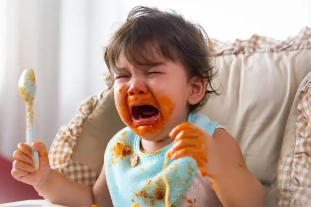 Photo of Adorable little toddler girl or infant baby crying when unsatisfied when finished eating food on baby chair. Cute infant girl get hungry and want more food. Mix race daughter get dirty Kid get tantrum