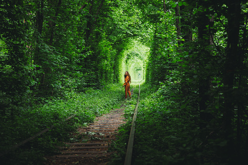 Young woman with long hair, in brown jumpsuit walking in the majestic abandoned natural green railway tunnel during summer sunrise - feeling awe and freedom