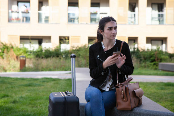 istock Female student with suitcase and bag sitting and using mobile phone 1256119247