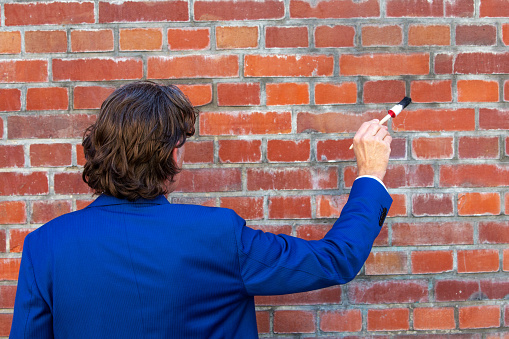 back of businessman in blue suit standing at brick wall with paintbrush