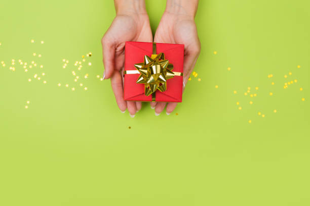 istock Women hands holding a gift or gift box decorated with confetti on a green table top view. 1256126362