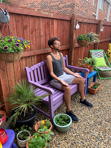 A side view of a young mixed-race gentleman sitting with his palms facing upwards in a yoga pose seated in his back garden relaxing in the north-east of England.