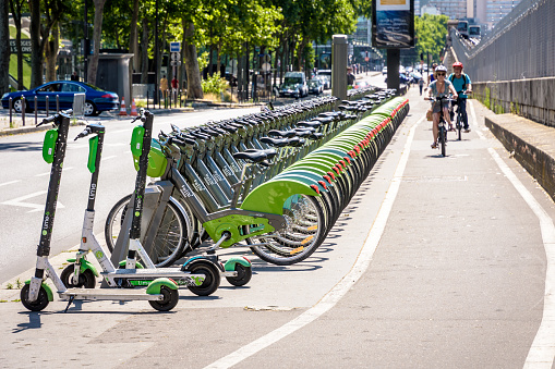 Paris, France - June 23, 2020: Cyclists are biking on a cycle lane past a fleet of Velib shared bicycles, lined up at the Velib docking station at Boulevard de Bercy along with Lime electric scooters.