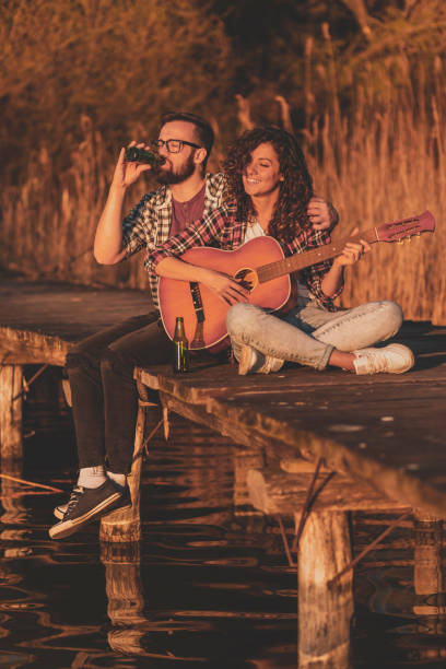 Couple sitting at the lake docks, playing the guitar, singing and drinking beer, enjoying a day in nature and relaxing