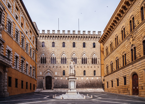 Siena, Tuscany, Italy - June 21, 2020: Monte dei Paschi di Siena headquarter. Empty place with no people. CLoudy sky. The headquarter of Monte dei Paschi di Siena bank is inside an historic building also called Rocca Salimbeni. At the center of the square stands the statue of Sallustio Bandini, a religious, political and Italian economist who lived in the eighteenth century, which is credited with the invention of the bill, while on the right is the Palazzo Spannocchia.