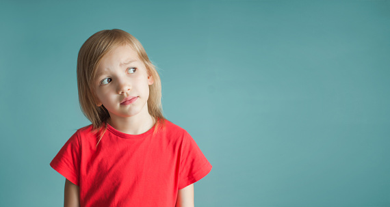 Close-up studio shot of a lovely little girl in a red t-shirt and posing against a blue background. Looks thoughtfully to the side. Copy space