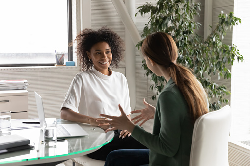 Happy diverse millennial women distracted from computer work chat talk in office during break, smiling young multiracial female colleagues have fun brainstorm discussing ideas or thoughts at workplace