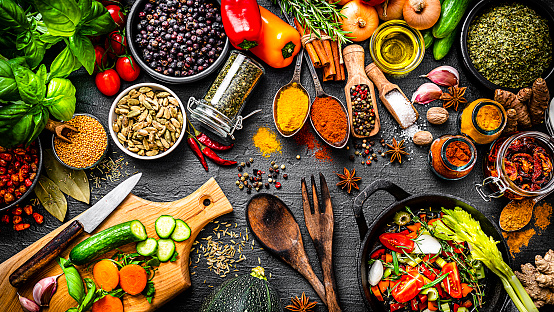 Cooking and seasoning backgrounds: large group of multicolored ingredients, spices and vegetables shot from above on black stone background. The composition includes spices like mustard seeds, paprika, turmeric, pepper, cinnamon sticks, star anise, cardamom, nutmeg, bay leaves, dried oregano and vegetables like onion, garlic, peppers, olive oil, rosemary, basil, dried tomatoes, cherry tomatoes, cucumber among others. A cutting board with vegetables and a cast iron pan complete the composition. High resolution 42Mp studio digital capture taken with SONY A7rII and Zeiss Batis 40mm F2.0 CF lens