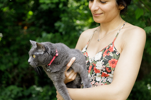 A woman holds a cat outside.  She is in a floral print dress and has olive skin and dark hair.  The cat has gray fur.  They are standing in front of some green foliage.  The woman is Iranian ethnicity, the cat is a Russian Blue.  She is smiling and looking towards the cat.