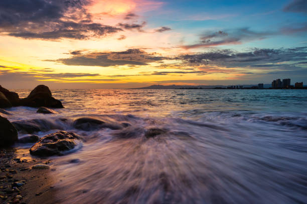 A Colorful Sunset on the Ocean With a Wave Rushing to Shore and Buildings in the Background