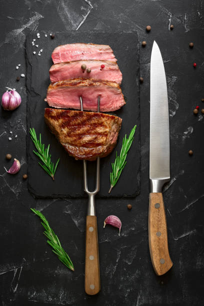 istock Grilled ribeye beef steak medium rare on the fork, black stone background. Top view, flat lay, copy space. 1256169131