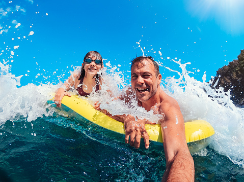 Father and daughter floating on yellow airbed and having fun on the beach while getting splashed by the wave