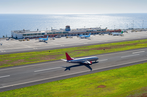 Aerial view of Funchal international airport with planes by terminal building, airplane taking off at air field, seascape in background in bright sunshine, Funchal, Madeira island, Portugal