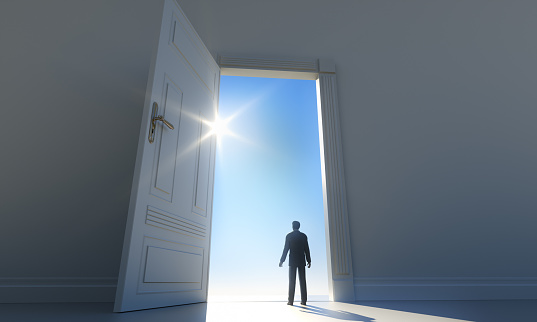 Man standing in a dark room with an open door to the sun with blue sky
