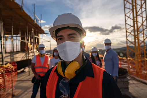 Portrait of a happy construction worker at a building site wearing a facemas with a group of workers at the background